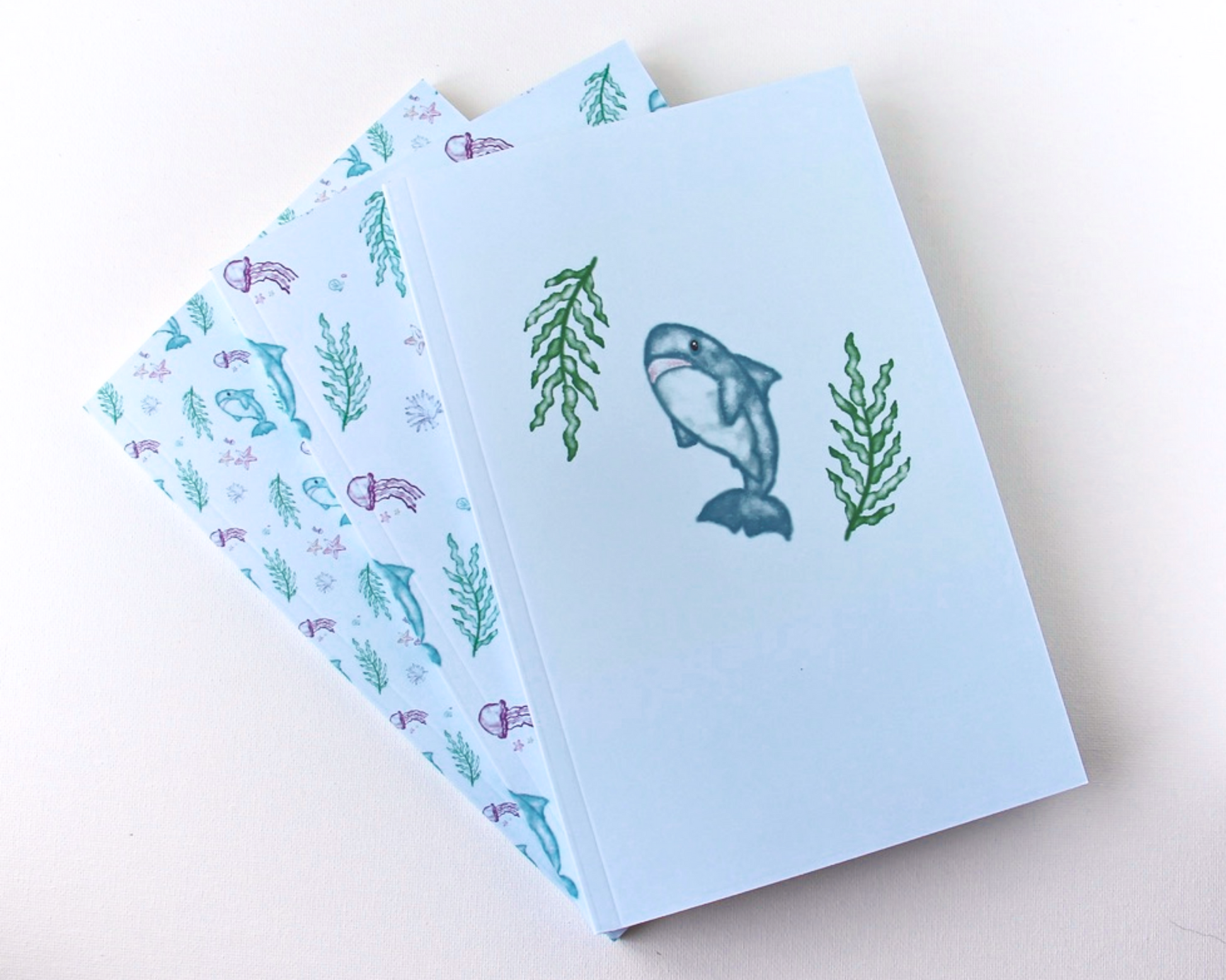 Set of three pastel blue notebooks spread out in a display, each with a watercolour pattern featuring sharks, jellyfish, starfish, shells, and seaweed.