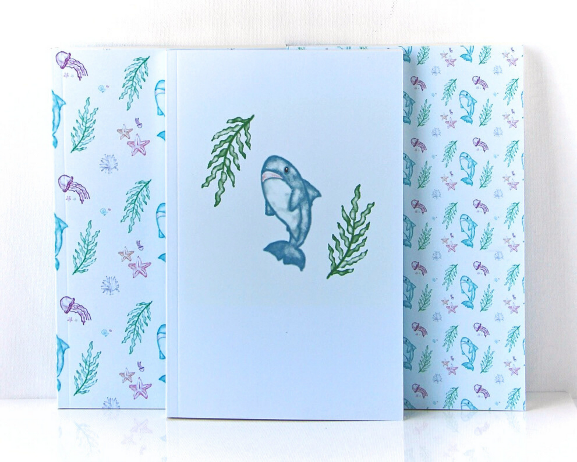 Set of three pastel blue notebooks spread out in a display, each with a watercolour pattern featuring sharks, jellyfish, starfish, shells, and seaweed.