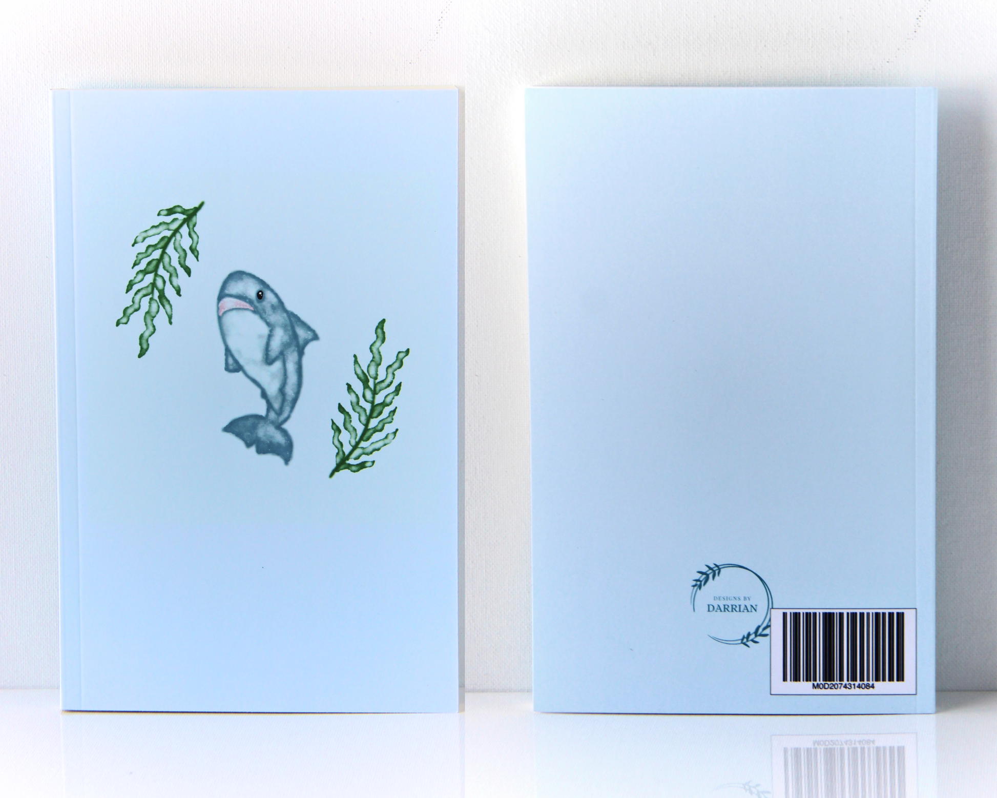 Pastel blue notebook with a watercolour shark and seaweed cover and a back side with a Designs By Darrian Logo on the bottom.