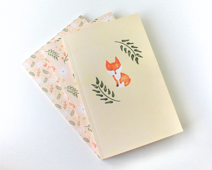 Notebook set of three spread out in a display with cream covers featuring a watercolour pattern with orange foxes, daisies, orange and yellow flowers, and leaves.