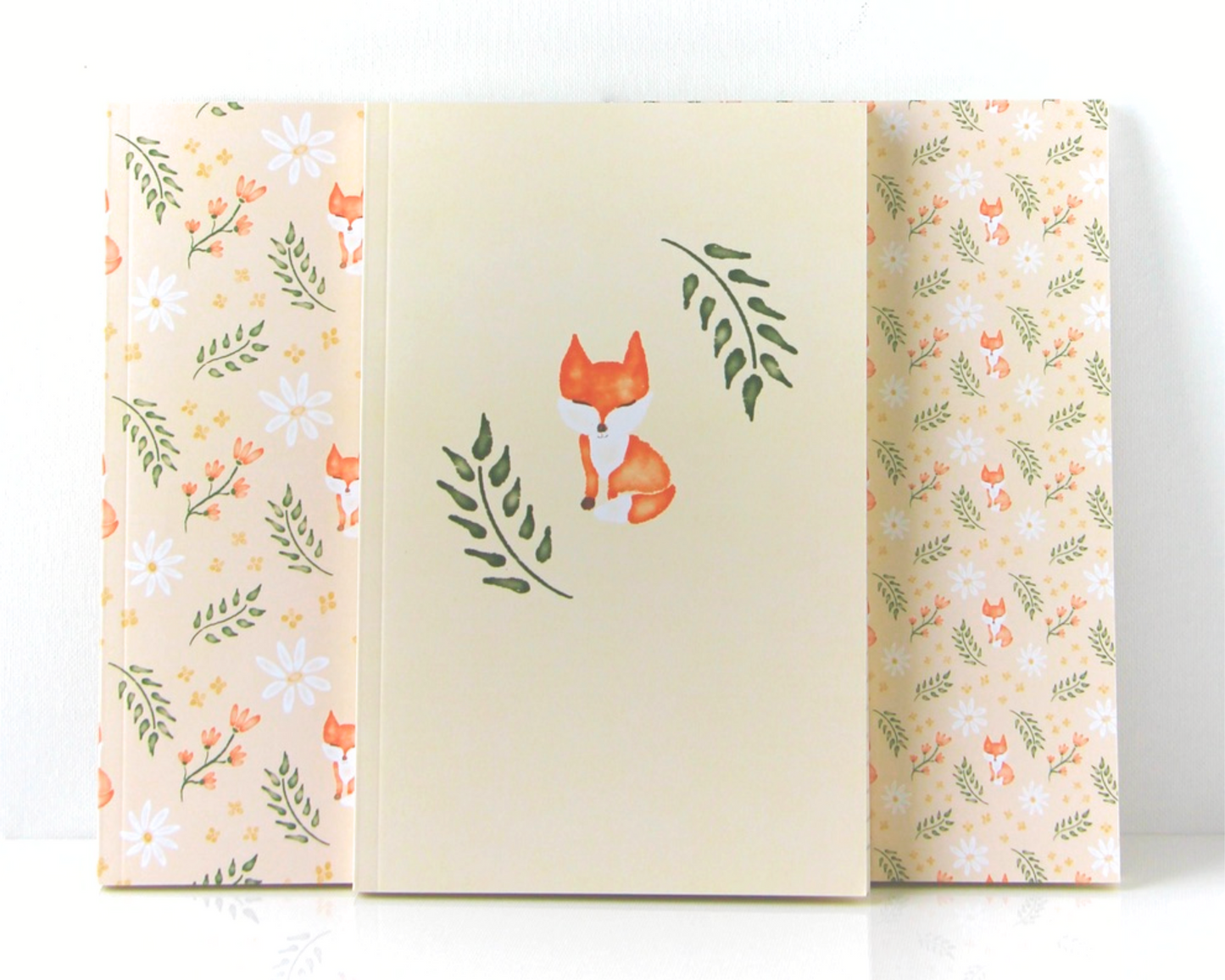 Notebook set of three spread out in a display with cream covers featuring a watercolour pattern with orange foxes, daisies, orange and yellow flowers, and leaves.