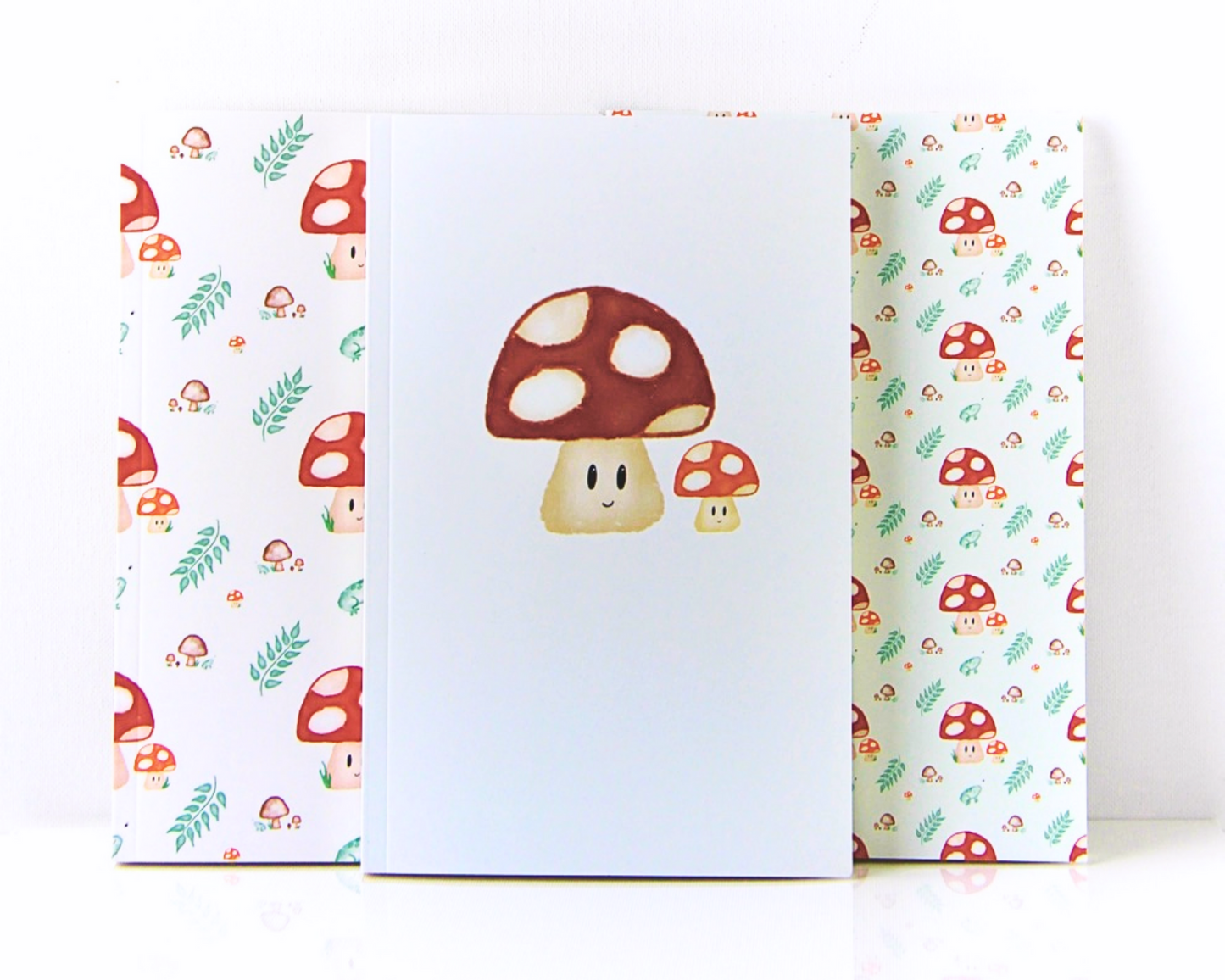 Notebook set of three spread out in a display, with a warm white background, with a watercolour pattern featuring red spotted mushrooms, leaves, brown mushrooms, and small green frogs.