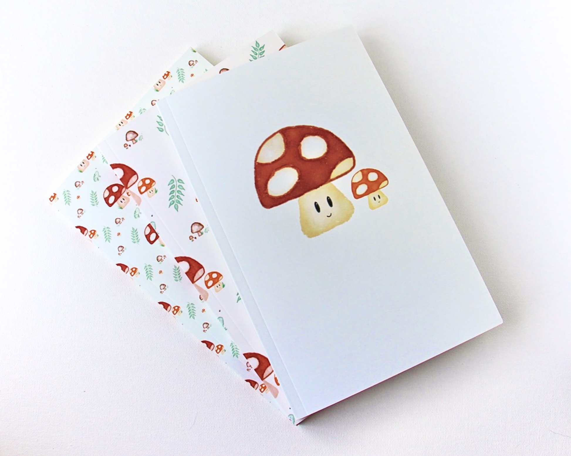 Notebook set of three spread out in a display, with a warm white background, with a watercolour pattern featuring red spotted mushrooms, leaves, brown mushrooms, and small green frogs.