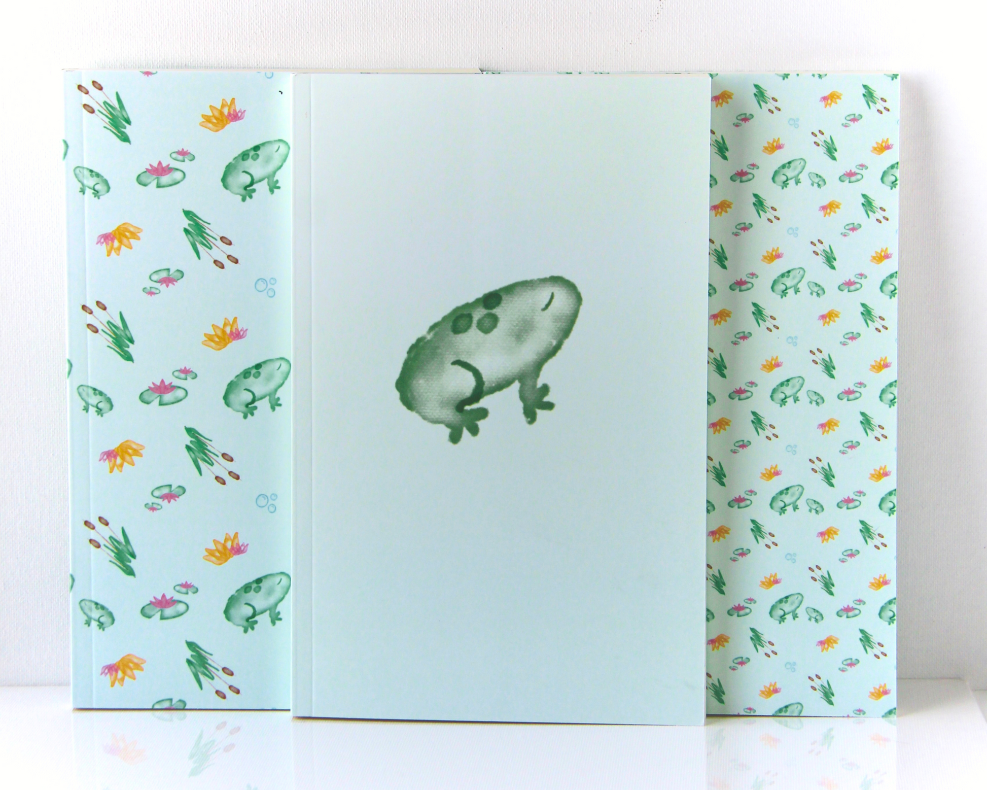 Pastel Green Notebook Set of Three display spread with covers featuring green watercolour frogs, cattails, lily pads, and orange and pink lotus flowers