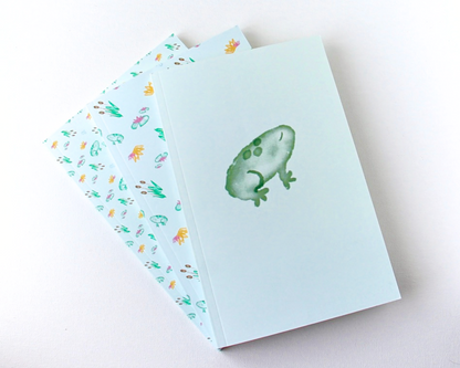 Pastel Green Notebook Set of Three display spread with covers featuring green watercolour frogs, cattails, lily pads, and orange and pink lotus flowers