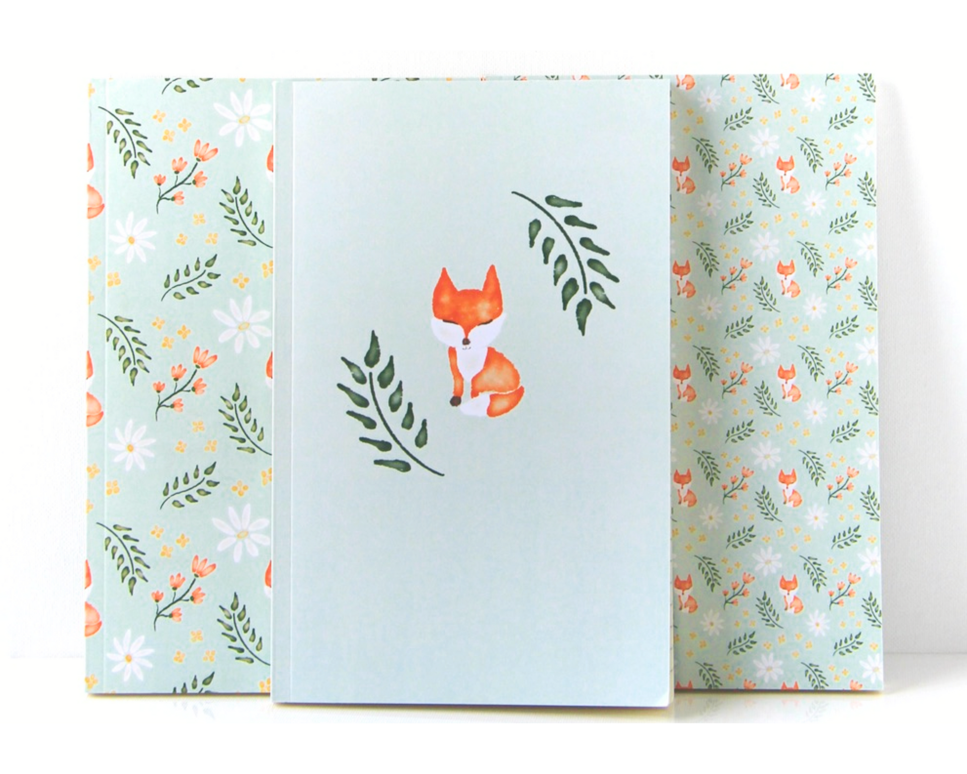 Notebook set of three spread out in a display with sage green covers featuring a watercolour pattern with orange foxes, daisies, orange and yellow flowers, and leaves.