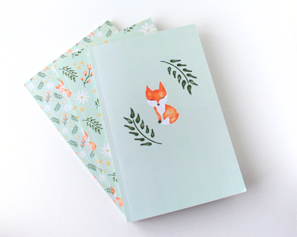 Notebook set of three spread out in a display with sage green covers featuring a watercolour pattern with orange foxes, daisies, orange and yellow flowers, and leaves.